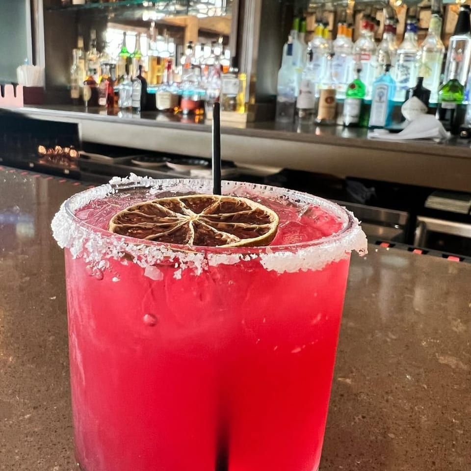 A vibrant red cocktail with a salted rim and dried citrus slice sits on a bar counter.