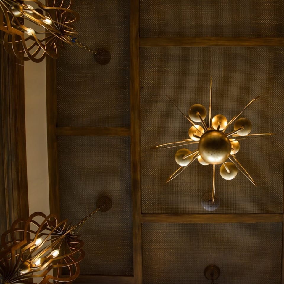 Ornate golden chandeliers hang from a dark, textured ceiling with wooden beams at Krinti Mediterranean Grill.